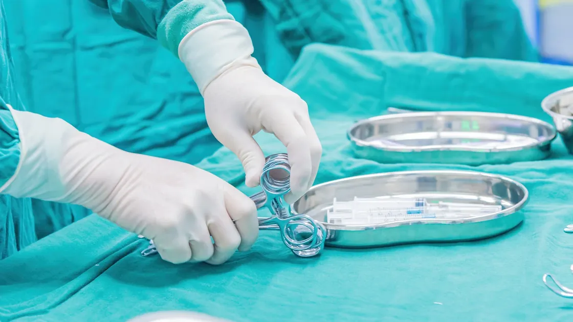 Gloved hands arrange metal medical instruments beside trays of syringes.