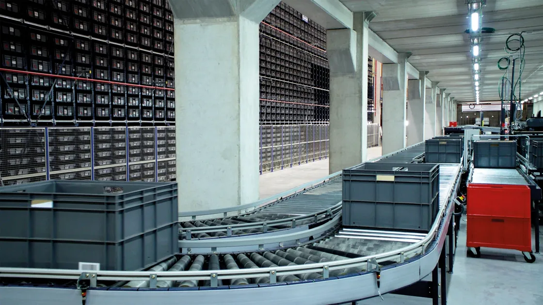Storage totes travel along a curved roller conveyor beside warehouse shelving.