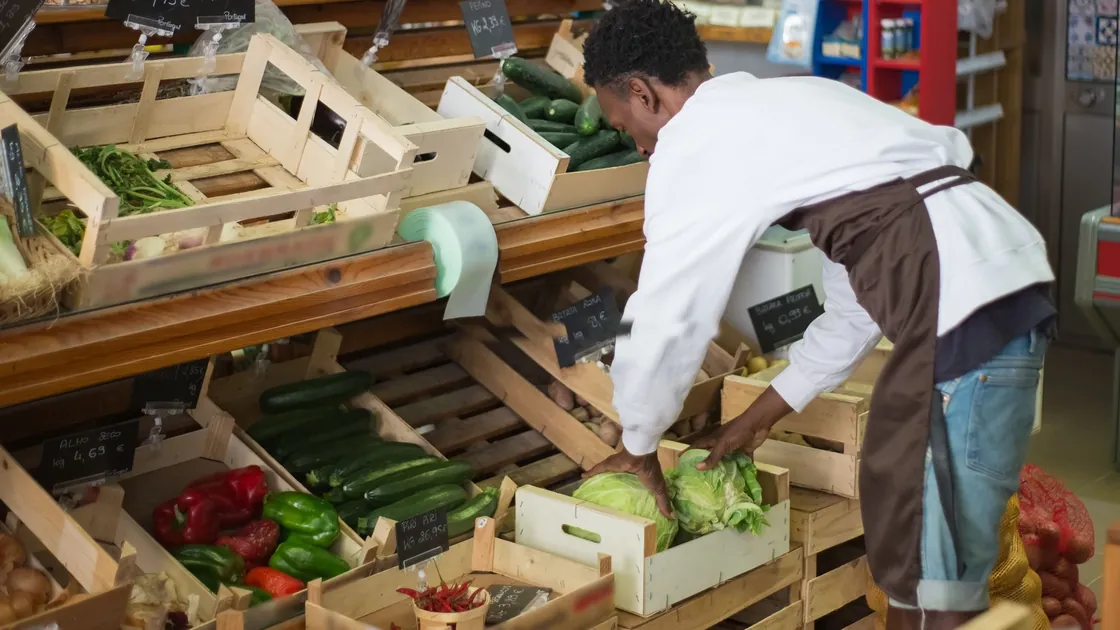Grocery worker replenishes fresh vegetables in wooden display crates.