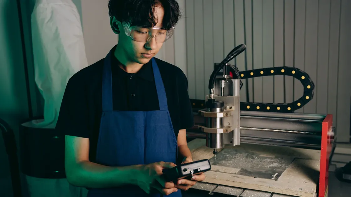 An engineer in safety glasses operates a handheld controller beside a CNC machine.