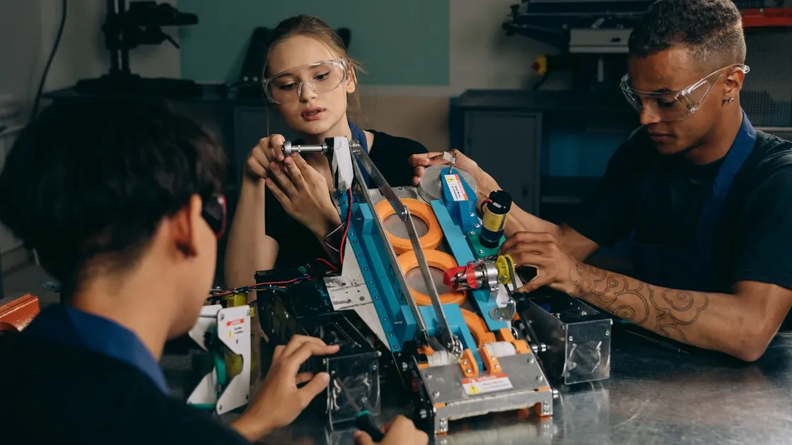 Three engineers in safety glasses work together on a robotic mechanism at a workbench.